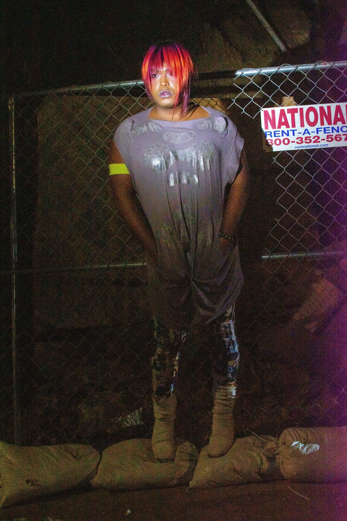 DJ Miss Parker posing with hands in their pockets against a fence illuminated by bright light